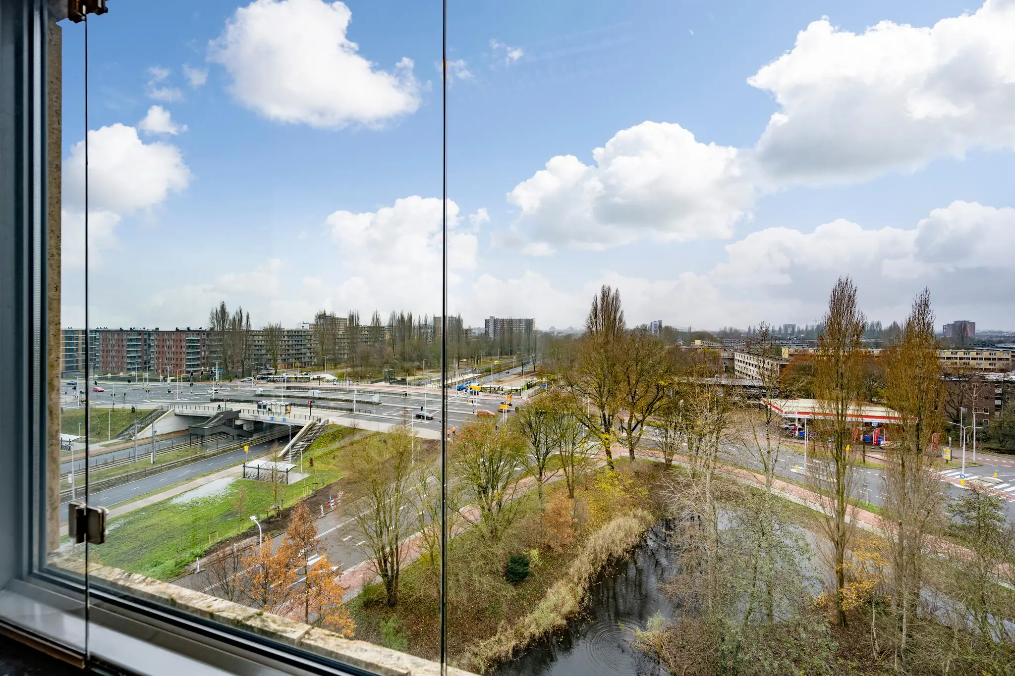 Uitzicht vanaf een hoger gelegen raam op de Burgemeester Rijnderslaan met verkeerswegen, een viaduct, een benzinestation en omliggende bebouwing.